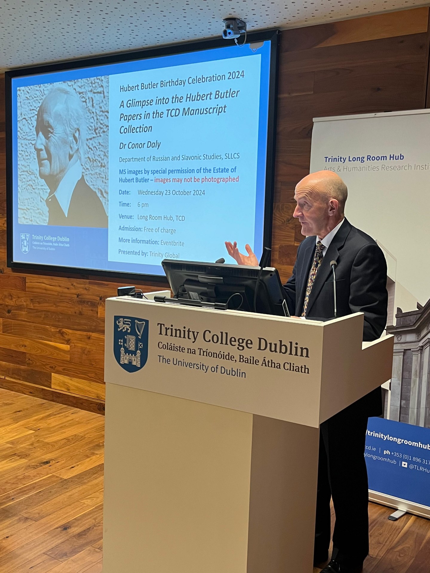 A man stands speaking at a pedastal with a screen behind him displaying the TCD logo