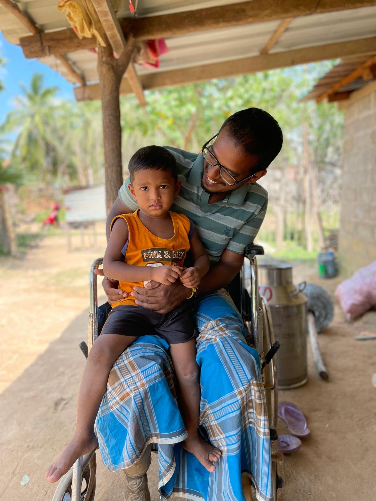 A man in a wheelchair holding a child on his lap. The child is looking directly to the camera with a serious look on his face, while the man's face is turned towards the child, smiling at him.